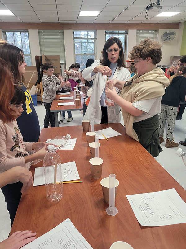 A woman in a white lab coat helps students with their science experiment which uses a clear plastic bottle.