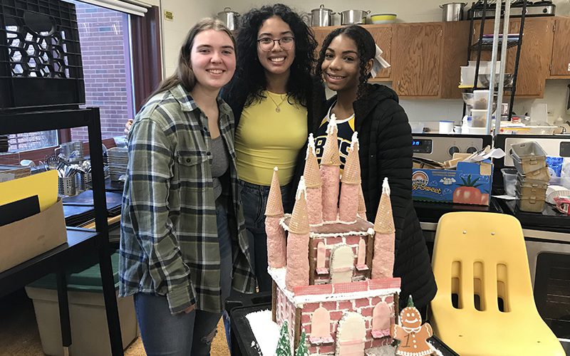 Three high school girls smile and stand behind a castle they made out of gingerbread.