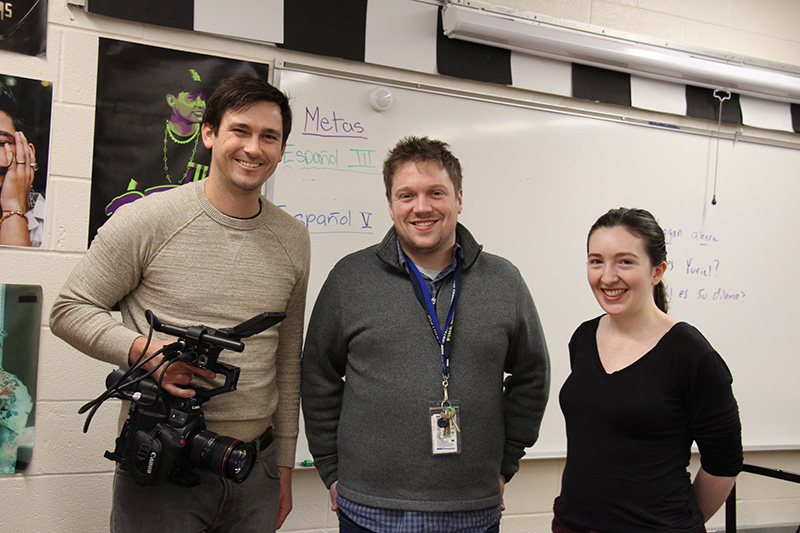 Three adults stand together in a classroom and smile. The man on the left is holding a video camera, has dark hair and is wearing a tan shirt The man in the middle has short dark hair and is smiling. The woman on the right has her long dark hair in a ponytail and is wearing a black shirt.