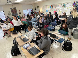 A crowded classroom with high school students sitting and standing around the perimeter.