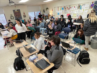 A crowded classroom with high school students sitting and standing around the perimeter.