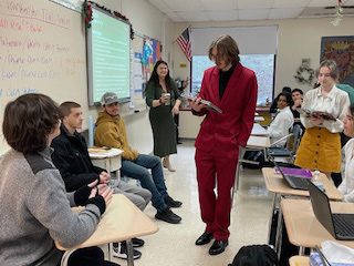 A high school student dressed in a maroon suit with a black shirt, reads to a group of other students sitting. Others are listening.