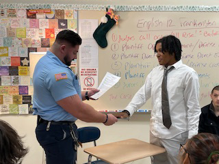 A police officer with short dark hair swears in a high school student for a mock trial. The student is dressed in a shirt and tie.