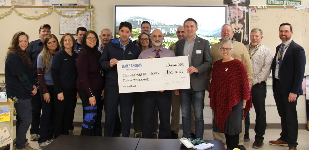 A group of 17 people stand in front of a large display screen. Three in the front hold a ceremonial check for $50,000.