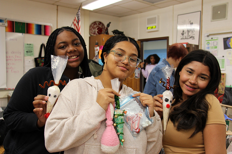 Three high school girls stand together and smile as they hold up their crafts.