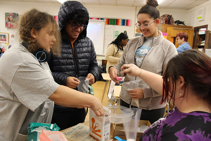 Four high school kids stand around a table and make crafts.