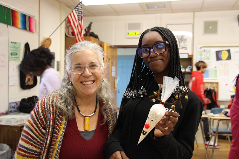 A woman with light hair smiles with a high school girl holding a craft.