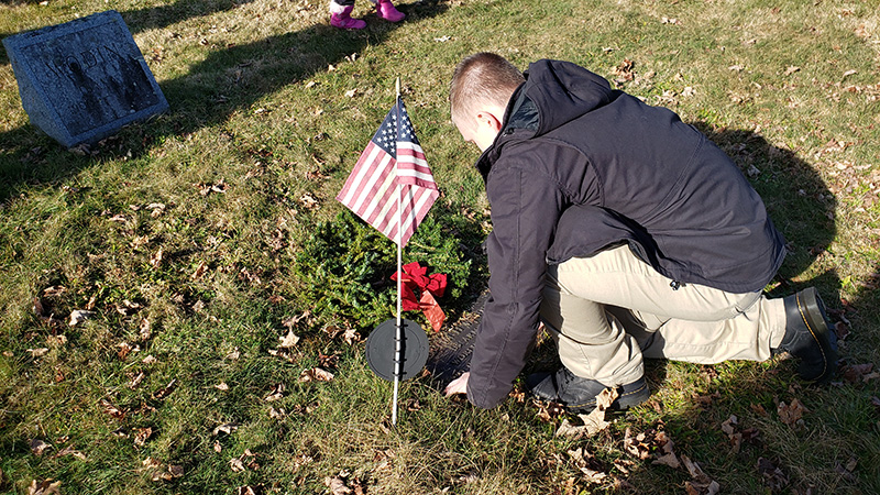A young man in a dark jacket and khaki pants bends over placing a wreath on a grave that also has an American flag.
