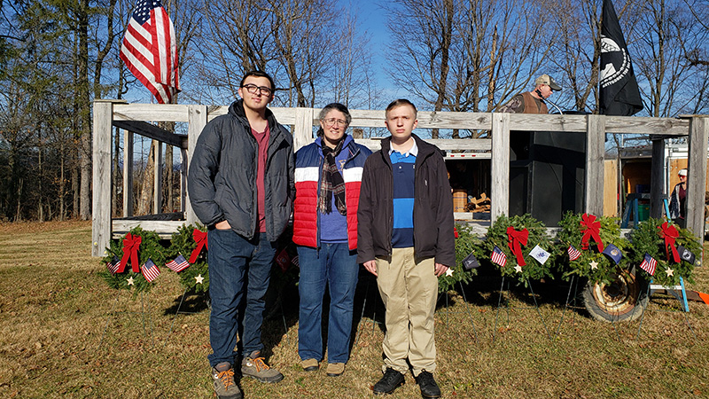 Two young men stand on either side of a woman. In the background is a monument with wreaths and an American flag.