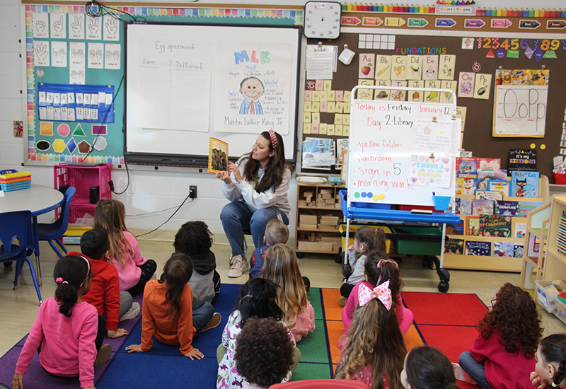 A group of pre-K students sit on a multi-colored rug while a women sits on a chair in the front holding up a book.