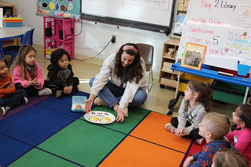 A woman with long dark hair sits cross-legged on a multi-colored rug with small kids on either side of her. They are looking at a white plate with two eggs on them.