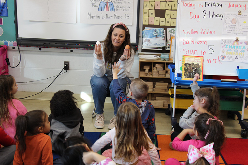 A woman with long dark hair sits in front of a group of pre-K students. She is holding up a white egg in one hand and a brown egg in the other.