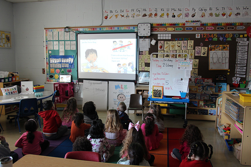 A group of about 18 pre-K students sit on a multi-colored rug in a darkened room watching a video book on a screen.