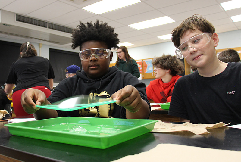 Two middle school boys sit together working on an experiment. The boy on the left lifts a green square out of the bubbles.