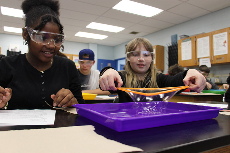 Two middle school girls with goggles lift a square out of bubble solution.