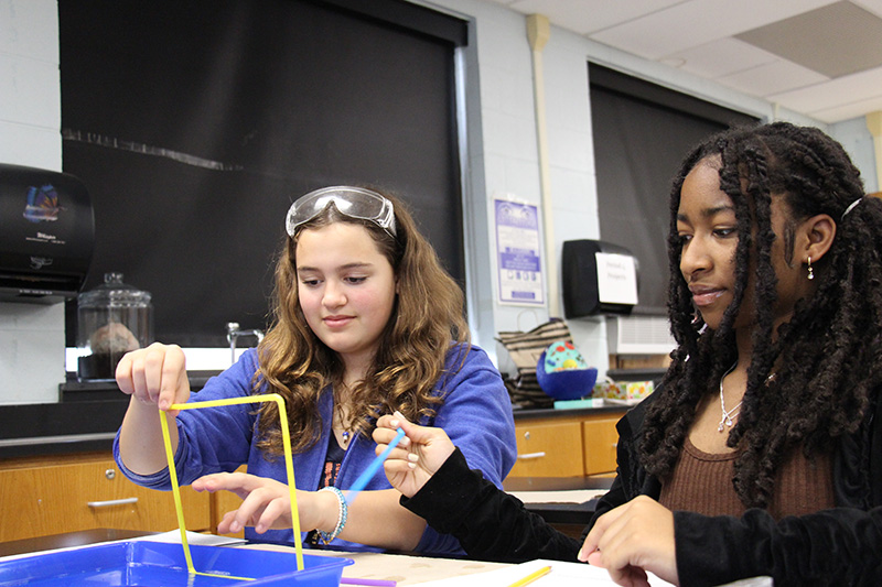 Two middle school girls hold up a large square with bubble membrane on it. One girl sticks her finger through it.