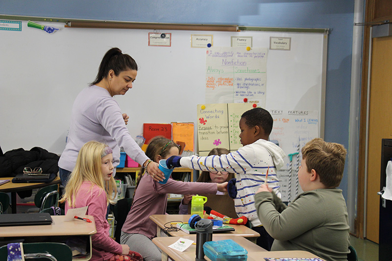 A classroom setting with third grad ekids working at their desks and a woman with dark hair in a ponytail offering a cup to one of the children.