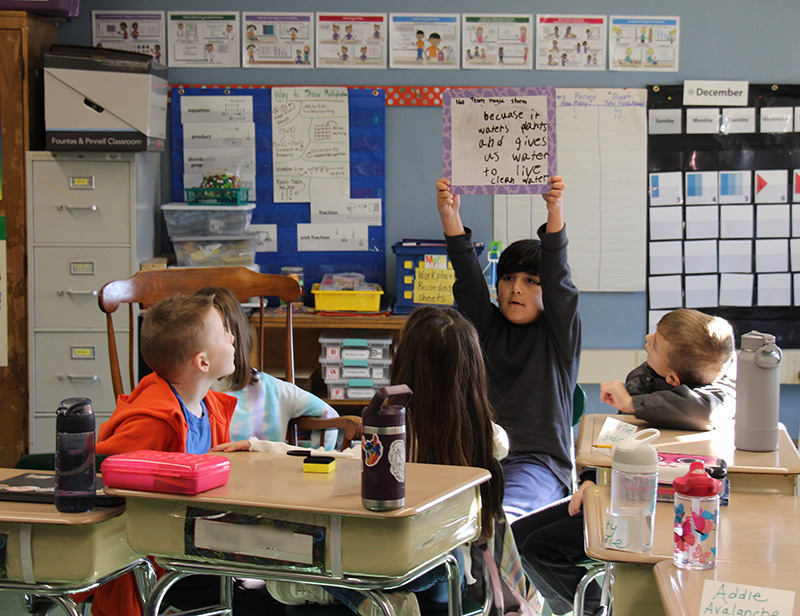 A classroom with a group of third grade students sitting at desks. One boy with short dark hair holds a whiteboard over his head.