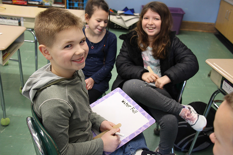 Three third graders sit in chairs in a semi circle and smile. A boy is holding a purple and white white board that says Waterfall on it. Two girls sign near him and smile as they work together.