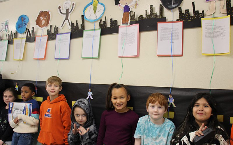 Seven fourth-graders stand by a wall by their classroom with their balloons above them and letters below the balloons. The kids are all smiling.