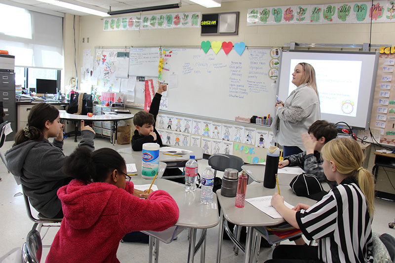 A woman at the front of the class teaches a class of fifth graders. One boy has his hand raised.