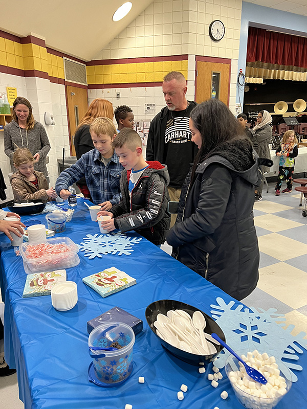 A group of kids and adults at a table with a blue table cloth and white snowflakes on it. They are getting cups of hot chocolate.
