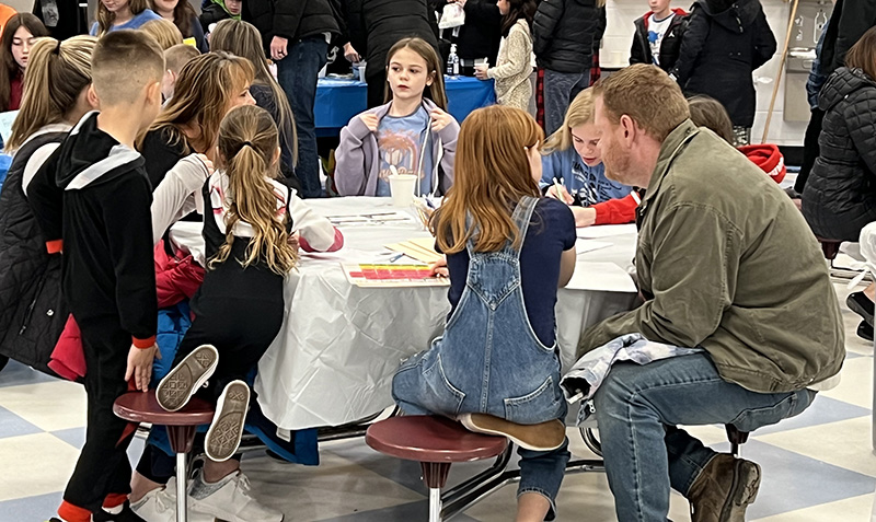 Elementary students and a couple adults sit around a table working.