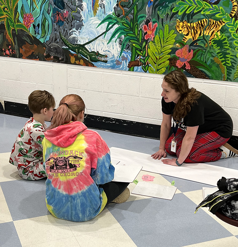 Two elementary students sit on the floor and work with an adult .
