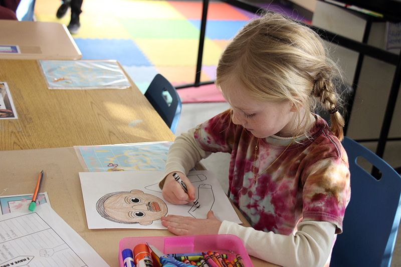 A girl with long blonde hair color a picture at her desk.