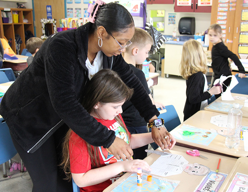 A woman with dark hair pulled back in a bun leans over a second-grade girl showing her how something is done on a piece of paper.