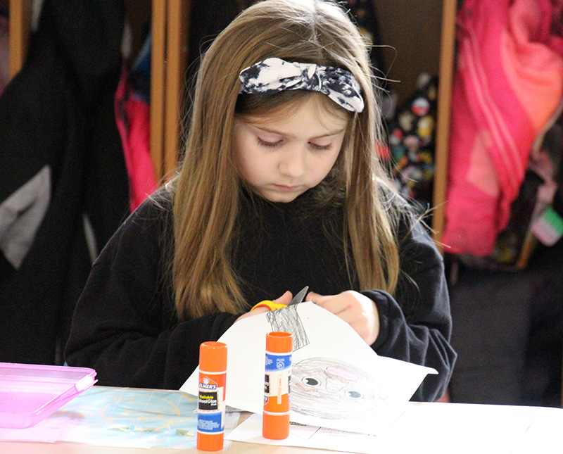 A second-grade girl with long brown hair with a black and white bow in it cuts a figure from a piece of paper.