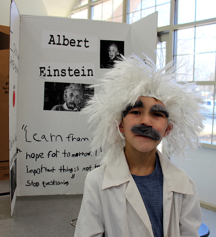 A fourth-grade boy dressed in a white lab coat with a big black mustache and crazy white hair stands in front of a poster board that says Albert Einstein with pictures of the scientist and information about him.