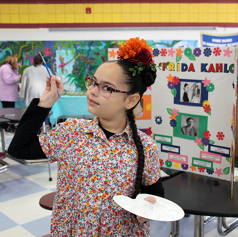 A girl with a long dark braid wearing a flowered shirt with flowers in her hair, holds a palate with paint on it and a paintbrush in her other hand.