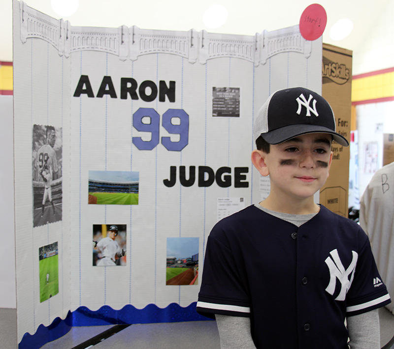 A boy dressed with a blue shirt with NY on it and a cap stands in front of a trifold poster that says Aaron Judge 99 with pictures an dinformation about him.