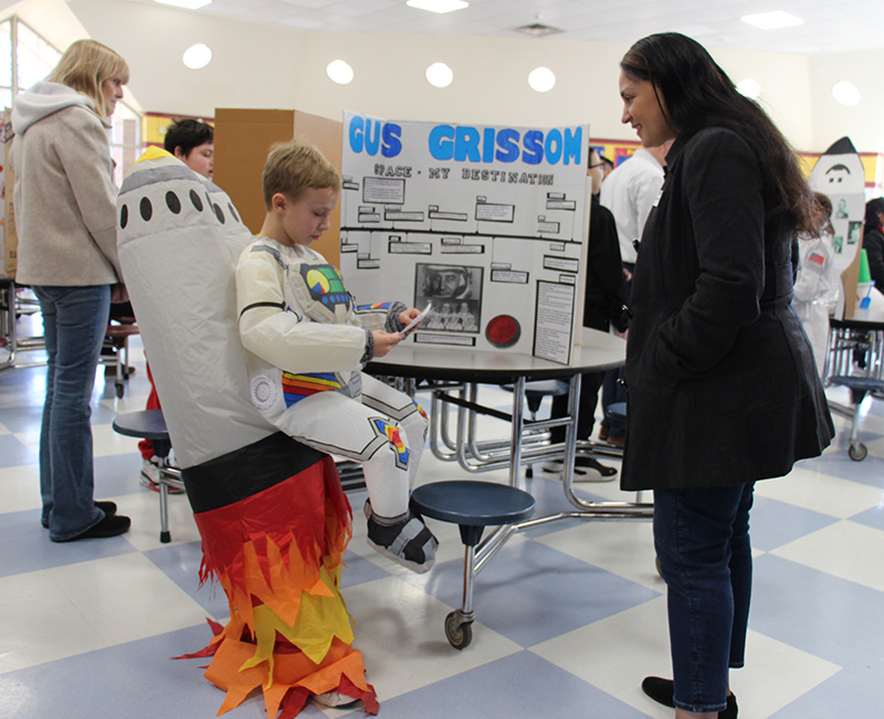 A fourth-grade boy wears an astronaut costume complete with a rocket on his back and orange ad red and yellow flames comin gout of the bottom. He is talking to a woman and they are standing in front of a board that has information about Gus Grissom on it.