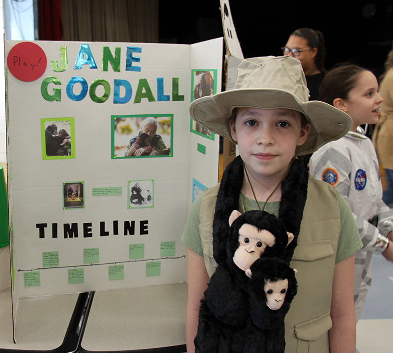 A fourth-grade girl wearing a safari hat and a stuffed monkey around her neck, stands in front of a poster that says Jane Goodall with information about her on it.