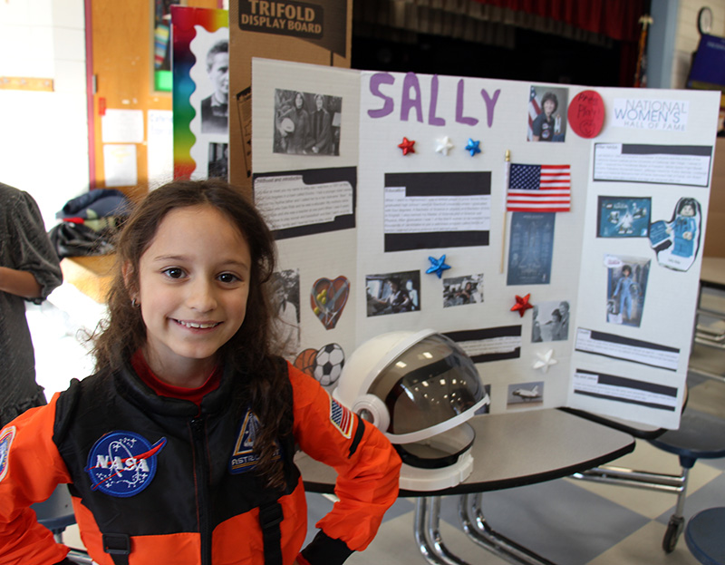 A fourth-grade girl smiles. She is wearing an orange and blue astronaut suit and stands in front of a poster about Sally Ride.