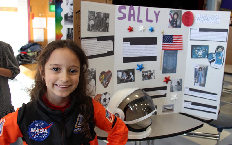 A fourth-grade girl, with long dark hair smiles. She is wearing an orange and black astronaut suit with a NASA patch on it. Behind her is a large trifold poster board with Sally Ride's pictures and information about the astronaut.