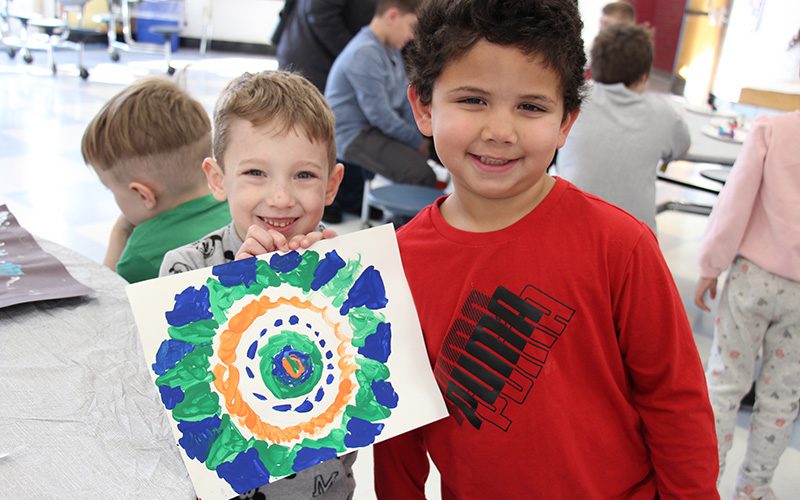Two elementary-age boys smile. The boy on left has short blonde hair and is holding up a painting of green and blue. The boy on the right has dark hair, is smiling and wearing a red shirt.