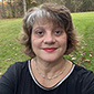 A woman with short brown hair smiles. She is wearing a black shirt and there is grass in the background.