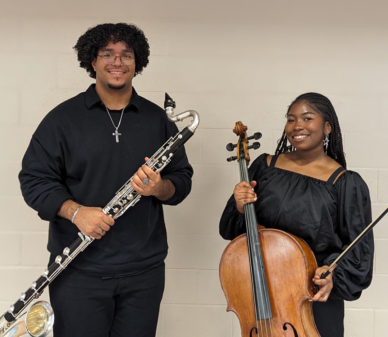 Two high school students stand and smile. The boy on the left is dressed in black and holds a large wind instrument. The young woman on the right is also dressed in black and holds a cello.