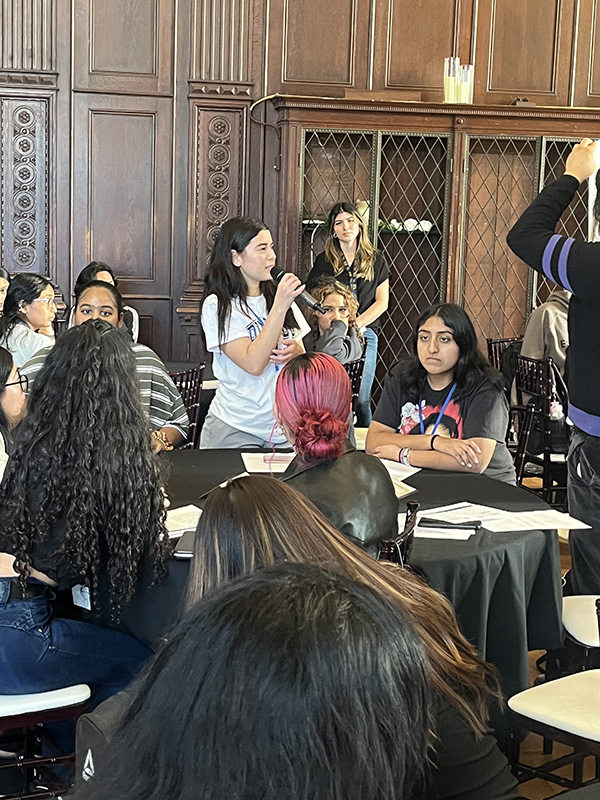 A room filled with high school students sitting at tables. One girl holds a microphone and is standing talking to the others.