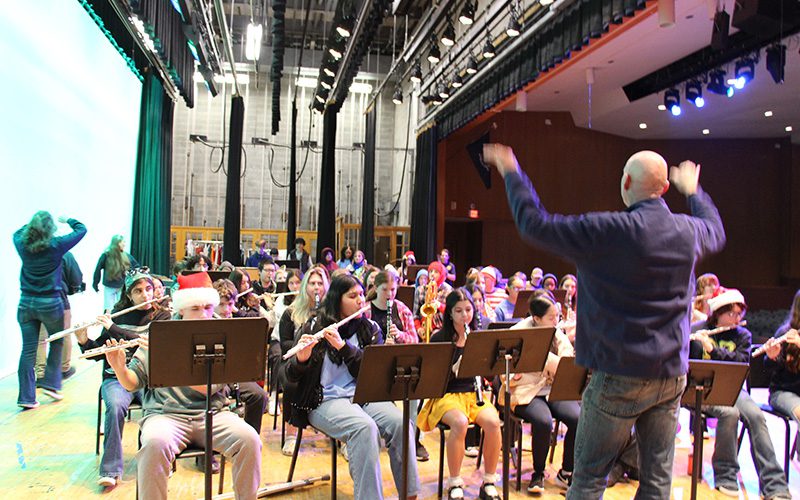 A large number of high school students sit on a stage and play instruments with a man in front conducting.