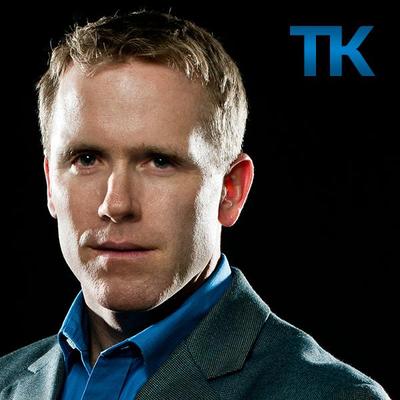 A black background with a TK in the upper right. A man with short light hair is pictured. He is wearing a blue shirt and suit jacket.