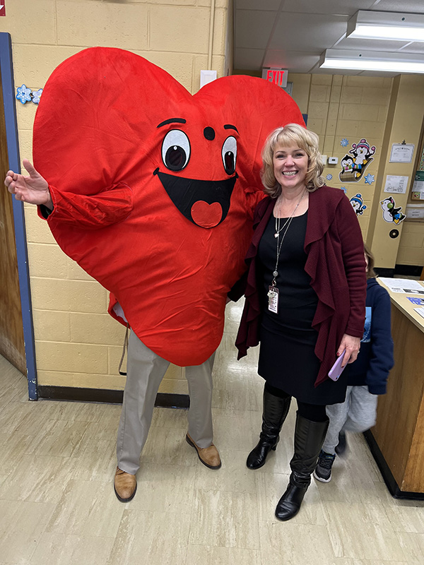 A woman with short blonde hair smiles as she poses with a person dressed in a big red heart costume.