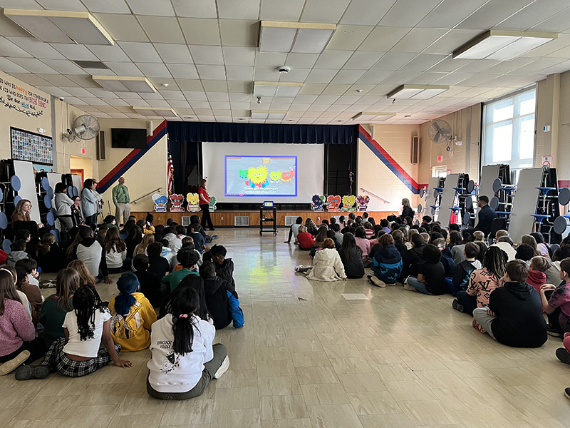 Two sides of a large room are filled with elementary students sitting on the floor. At the front is a large screen and a woman talking about heart health.