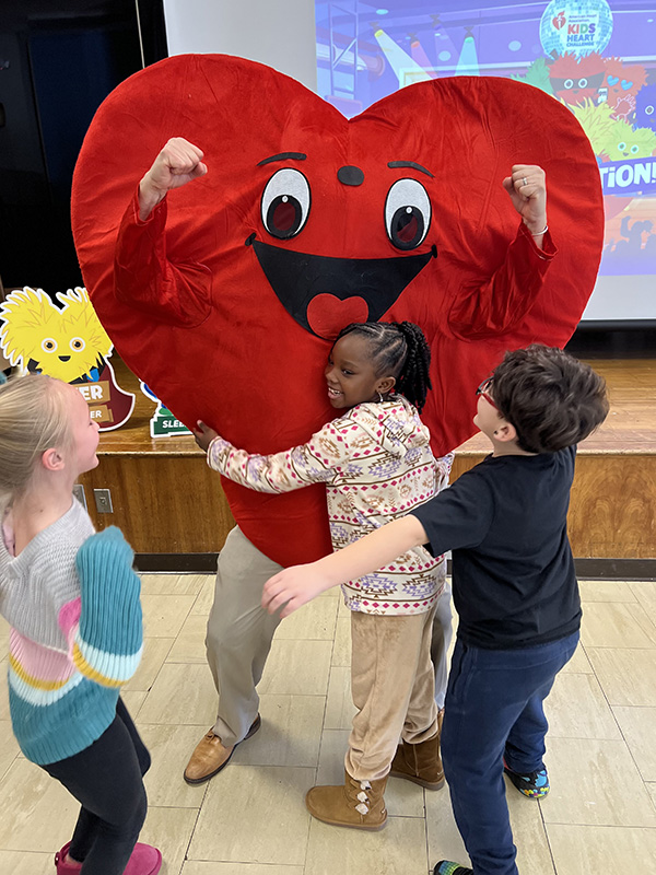Young elementary students hug a person who is dressed in a big red heart costume.