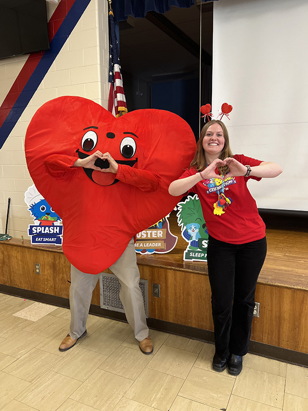 A woman stands on the right making a heart sign with her hands and she has a headband on with two bouncy hearts on it. On the left is a person dressed in a big red heart costume, also making a heart with his hands.