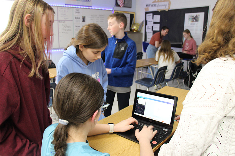 A middle school girl sits working on a Chromebook while three other girls and one boy stand around her. They are all working on a flyer.