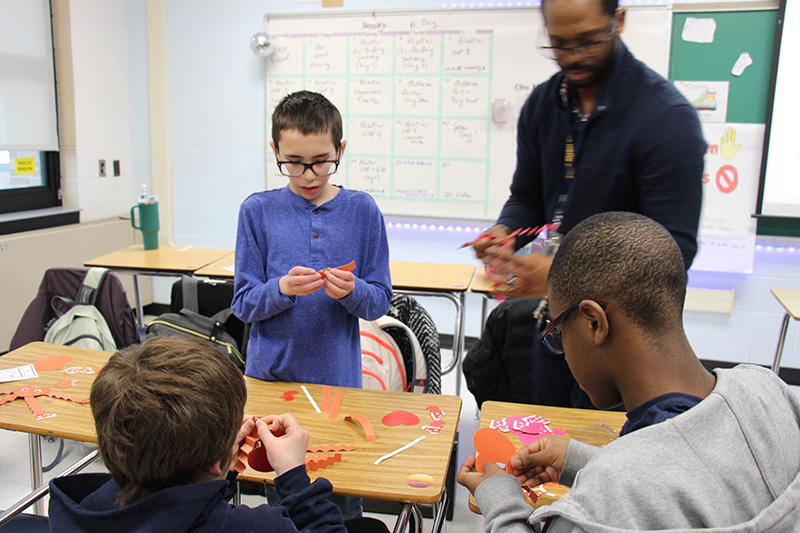 A man with glasses stands by as three students are making valentines with heart cut outs and folded paper for legs and arms.